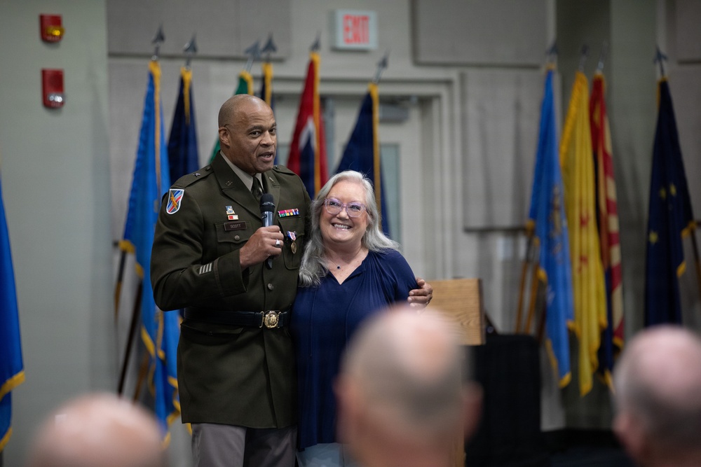 A U.S. Army officer in dress uniform holds a microphone and stands alongside a civilian woman with gray hair and glasses at what appears to be a military ceremony or retirement event. The officer has his arm around the woman and both are smiling toward an audience. Multiple military and state flags are displayed in the background.