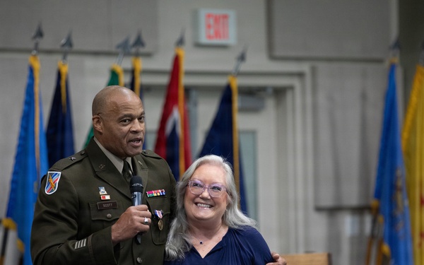 A U.S. Army officer in dress uniform holds a microphone and stands alongside a civilian woman with gray hair and glasses at what appears to be a military ceremony or retirement event. The officer has his arm around the woman and both are smiling toward an audience. Multiple military and state flags are displayed in the background.