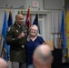 A U.S. Army officer in dress uniform holds a microphone and stands alongside a civilian woman with gray hair and glasses at what appears to be a military ceremony or retirement event. The officer has his arm around the woman and both are smiling toward an audience. Multiple military and state flags are displayed in the background.