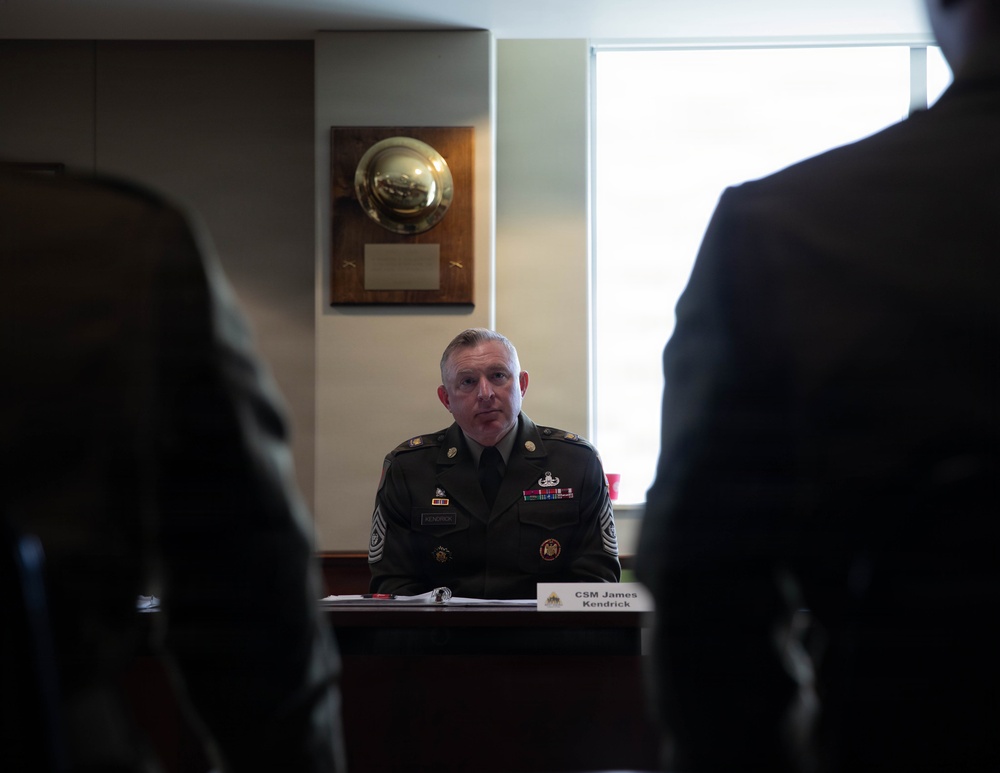A U.S. Army Command Sergeant Major named James Kendrick is seated at a desk or conference table in what appears to be an official military meeting or briefing room. He is wearing the Army Service Uniform (ASU) with visible rank insignia, ribbons, and badges. A wooden award plaque is mounted on the wall behind him, and a name placard reading 'CSM James Kendrick' is visible on the table in front of him.