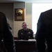 A U.S. Army Command Sergeant Major named James Kendrick is seated at a desk or conference table in what appears to be an official military meeting or briefing room. He is wearing the Army Service Uniform (ASU) with visible rank insignia, ribbons, and badges. A wooden award plaque is mounted on the wall behind him, and a name placard reading 'CSM James Kendrick' is visible on the table in front of him.