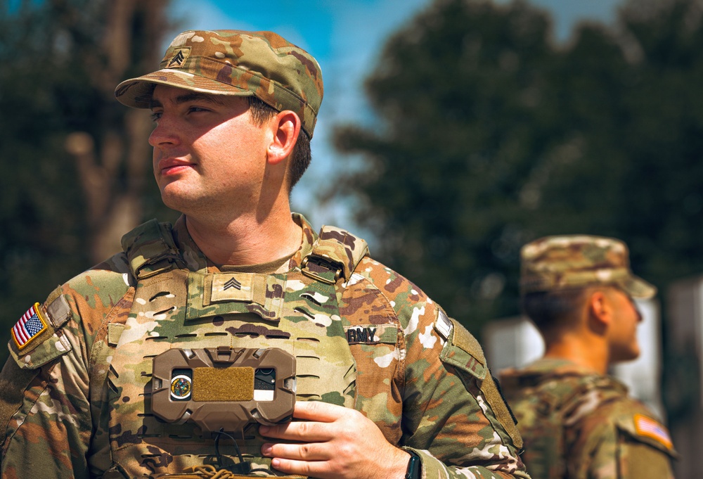 A U.S. Army Sergeant in the foreground is standing at attention, wearing Operational Camouflage Pattern (OCP) uniform with body armor and holding a device to his chest. A second soldier in matching OCP uniform is visible in the background, slightly out of focus. The setting appears to be an outdoor military formation or ceremony.