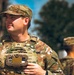 A U.S. Army Sergeant in the foreground is standing at attention, wearing Operational Camouflage Pattern (OCP) uniform with body armor and holding a device to his chest. A second soldier in matching OCP uniform is visible in the background, slightly out of focus. The setting appears to be an outdoor military formation or ceremony.