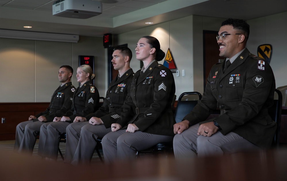 Five U.S. Army soldiers in Army Service Uniforms (ASUs) are seated in a row during what appears to be a formal military ceremony or event. They are wearing the olive green Army Service Uniform with various ribbons, badges, and rank insignia visible. The setting appears to be an official military briefing or conference room, with unit insignia patches visible on the wall behind them.