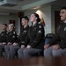 Five U.S. Army soldiers in Army Service Uniforms (ASUs) are seated in a row during what appears to be a formal military ceremony or event. They are wearing the olive green Army Service Uniform with various ribbons, badges, and rank insignia visible. The setting appears to be an official military briefing or conference room, with unit insignia patches visible on the wall behind them.