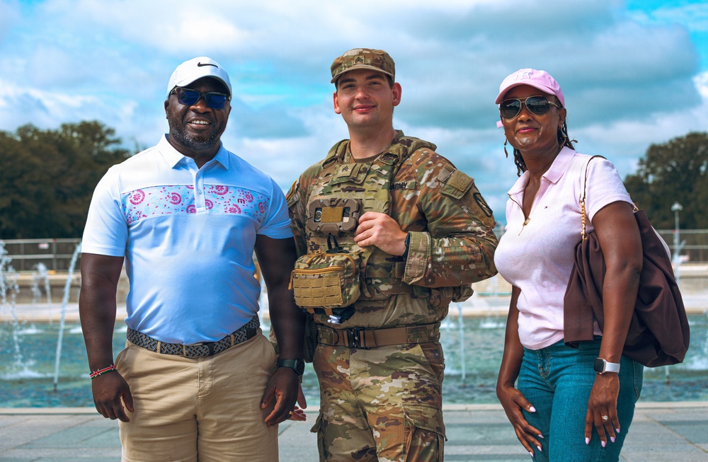 Three individuals pose together outdoors in front of a fountain on a partly cloudy day. The central figure is wearing U.S. Army military uniform with camouflage pattern and a patrol cap, flanked by two civilians dressed in casual attire. The setting appears to be a public landmark or memorial area.
