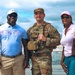 Three individuals pose together outdoors in front of a fountain on a partly cloudy day. The central figure is wearing U.S. Army military uniform with camouflage pattern and a patrol cap, flanked by two civilians dressed in casual attire. The setting appears to be a public landmark or memorial area.