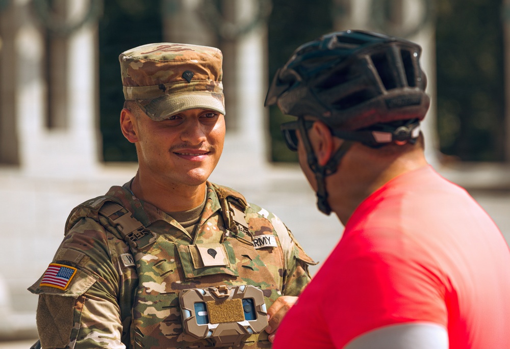 A smiling U.S. Army soldier in full OCP (Operational Camouflage Pattern) uniform is engaged in a friendly conversation with a civilian wearing a red shirt and black bicycle helmet. The soldier is wearing a body camera or electronic device on their chest rig and has a U.S. flag patch on their left shoulder. The setting appears to be an outdoor public area with classical architecture visible in the blurred background.