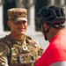 A smiling U.S. Army soldier in full OCP (Operational Camouflage Pattern) uniform is engaged in a friendly conversation with a civilian wearing a red shirt and black bicycle helmet. The soldier is wearing a body camera or electronic device on their chest rig and has a U.S. flag patch on their left shoulder. The setting appears to be an outdoor public area with classical architecture visible in the blurred background.