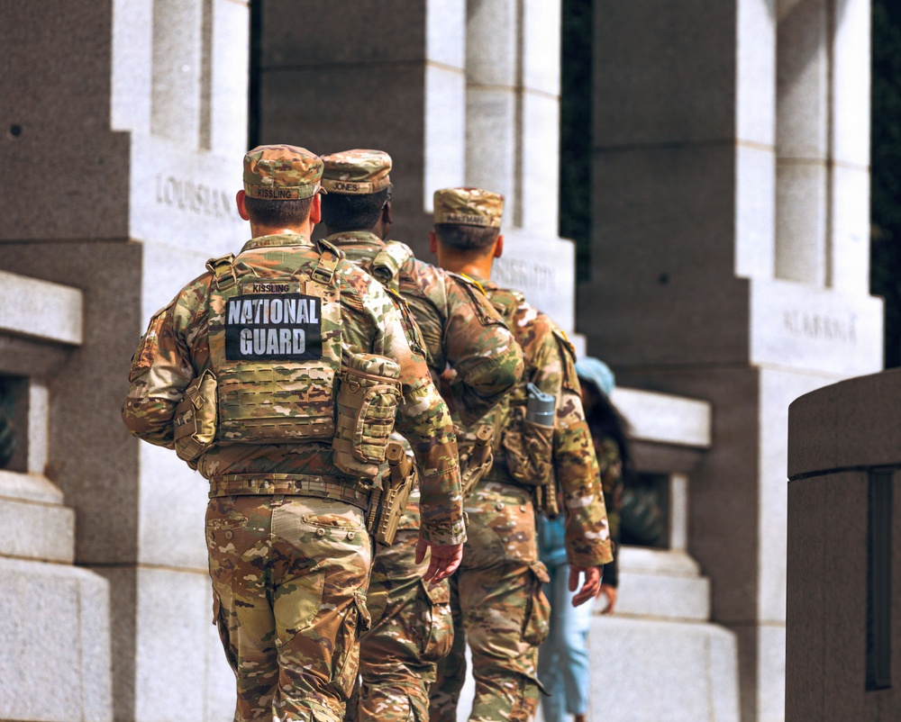 Three National Guard soldiers in multicam camouflage uniforms are walking away from the camera toward a large granite memorial structure. The leftmost soldier has a clearly visible 'NATIONAL GUARD' patch on their tactical vest with the name tag 'KISSLING'. The setting appears to be the National World War II Memorial in Washington, D.C., identifiable by the granite pillars with state names inscribed, including 'LOUISIANA' visible in the background.