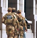 Three National Guard soldiers in multicam camouflage uniforms are walking away from the camera toward a large granite memorial structure. The leftmost soldier has a clearly visible 'NATIONAL GUARD' patch on their tactical vest with the name tag 'KISSLING'. The setting appears to be the National World War II Memorial in Washington, D.C., identifiable by the granite pillars with state names inscribed, including 'LOUISIANA' visible in the background.