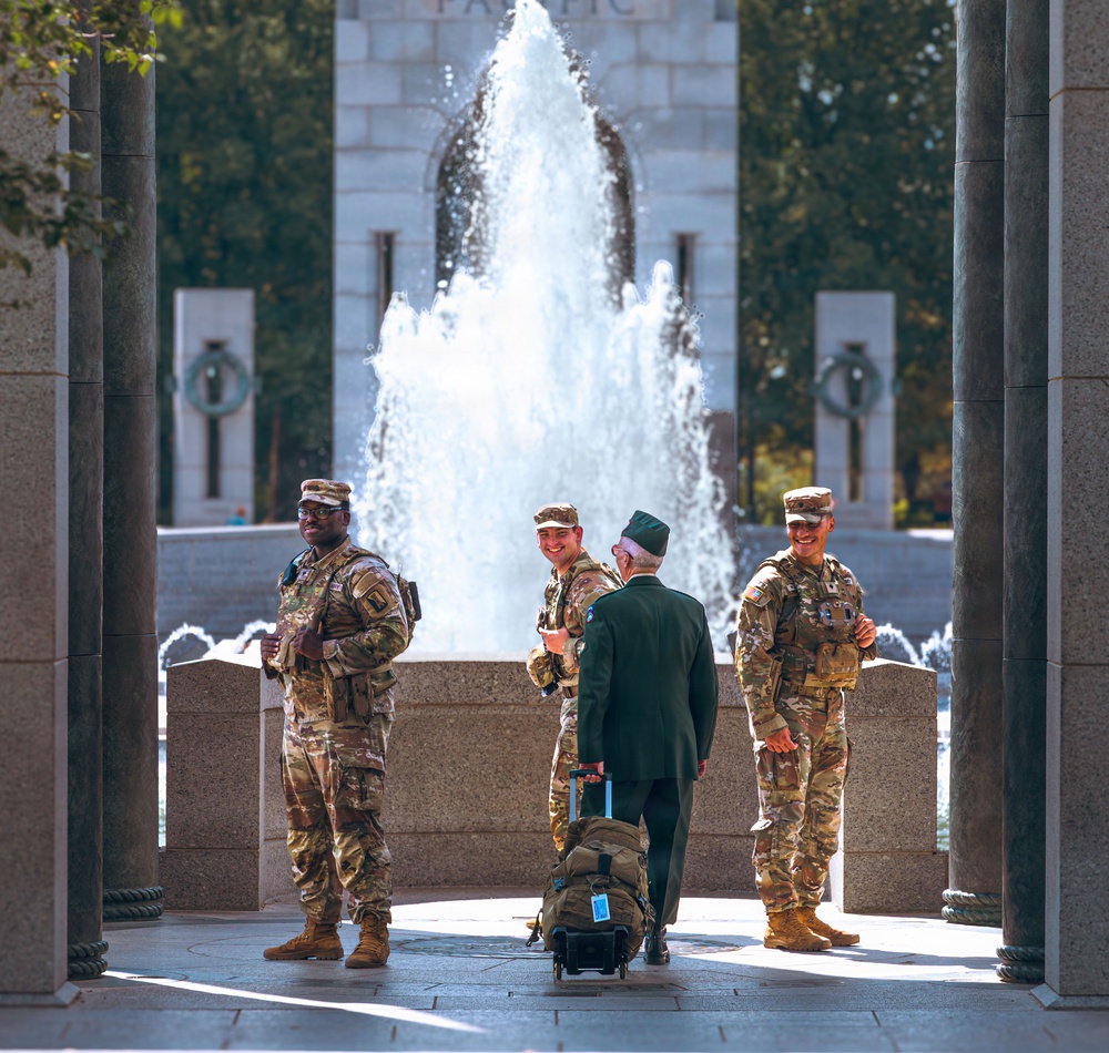 Three U.S. Army soldiers in modern OCP (Operational Camouflage Pattern) uniforms are interacting with an elderly veteran dressed in a dark military-style uniform at what appears to be the National World War II Memorial in Washington, D.C. The veteran, facing away from the camera and pulling wheeled luggage with a backpack, is engaged in conversation with the smiling soldiers. A large fountain and the memorial's iconic granite pillars and arches are prominently visible in the background.