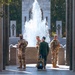 Three U.S. Army soldiers in modern OCP (Operational Camouflage Pattern) uniforms are interacting with an elderly veteran dressed in a dark military-style uniform at what appears to be the National World War II Memorial in Washington, D.C. The veteran, facing away from the camera and pulling wheeled luggage with a backpack, is engaged in conversation with the smiling soldiers. A large fountain and the memorial's iconic granite pillars and arches are prominently visible in the background.