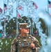 A U.S. Army soldier in MultiCam (OCP) camouflage uniform stands at attention or in a relaxed pose surrounded by multiple American flags on flagpoles. The image is shot through what appears to be a water fountain or bubble effect in the foreground, creating a bokeh-like visual distortion. The setting appears to be an outdoor public event or ceremony with spectators visible in the background.