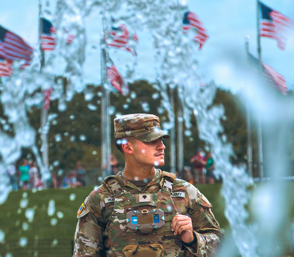 A U.S. Army soldier in MultiCam (OCP) camouflage uniform stands at attention or in a relaxed pose surrounded by multiple American flags on flagpoles. The image is shot through what appears to be a water fountain or bubble effect in the foreground, creating a bokeh-like visual distortion. The setting appears to be an outdoor public event or ceremony with spectators visible in the background.