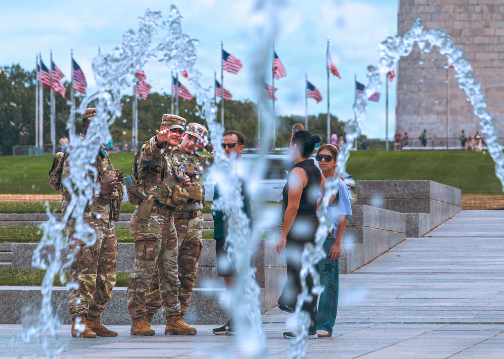 The image shows military personnel in camouflage uniforms and civilians gathered near a fountain at what appears to be the National Mall in Washington, D.C. Multiple American flags are visible on flagpoles in the background, along with a large stone monument structure on the right. The foreground features water jets from a fountain that partially obscure the subjects.