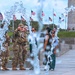 The image shows military personnel in camouflage uniforms and civilians gathered near a fountain at what appears to be the National Mall in Washington, D.C. Multiple American flags are visible on flagpoles in the background, along with a large stone monument structure on the right. The foreground features water jets from a fountain that partially obscure the subjects.