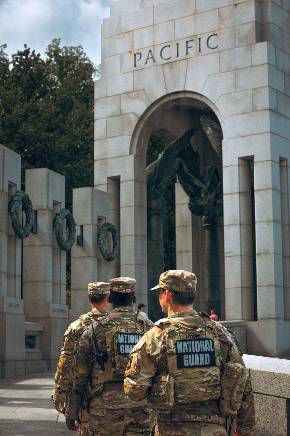 Three U.S. National Guard soldiers in MultiCam camouflage uniforms stand facing the Pacific Arch of the National World War II Memorial in Washington, D.C. The soldiers are viewed from behind, with 'NATIONAL GUARD' patches clearly visible on their backs and name tapes on their caps. Bronze wreaths and an eagle sculpture are visible within the memorial's archway.