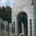 Three U.S. National Guard soldiers in MultiCam camouflage uniforms stand facing the Pacific Arch of the National World War II Memorial in Washington, D.C. The soldiers are viewed from behind, with 'NATIONAL GUARD' patches clearly visible on their backs and name tapes on their caps. Bronze wreaths and an eagle sculpture are visible within the memorial's archway.