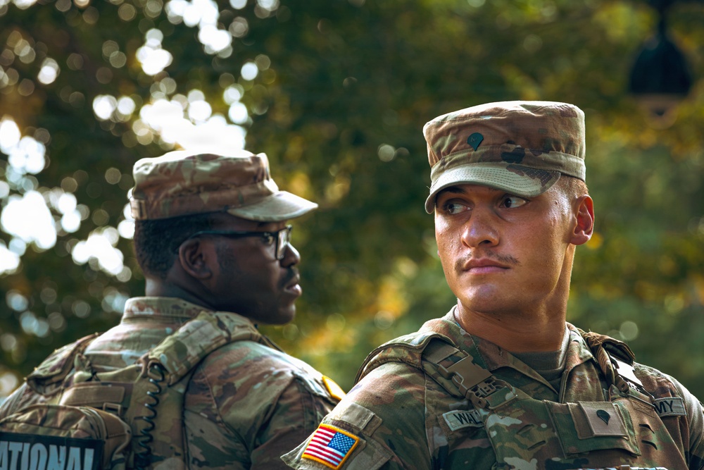 Two U.S. Army soldiers in MultiCam (OCP) camouflage uniforms are photographed outdoors against a bokeh green tree background. The soldier in the foreground faces right with a focused expression, while the soldier in the background faces left wearing glasses. Both are in full uniform with patrol caps and tactical gear.