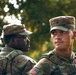Two U.S. Army soldiers in MultiCam (OCP) camouflage uniforms are photographed outdoors against a bokeh green tree background. The soldier in the foreground faces right with a focused expression, while the soldier in the background faces left wearing glasses. Both are in full uniform with patrol caps and tactical gear.