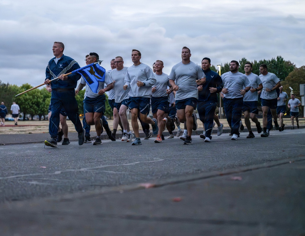 A large group of U.S. Air Force personnel are conducting a formation run on a paved road, likely on a military base. A lead runner on the left carries a blue unit guidon/pennant on a wooden staff. The group is dressed in standard PT (physical training) uniforms consisting of gray t-shirts and navy blue shorts.