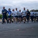 A large group of U.S. Air Force personnel are conducting a formation run on a paved road, likely on a military base. A lead runner on the left carries a blue unit guidon/pennant on a wooden staff. The group is dressed in standard PT (physical training) uniforms consisting of gray t-shirts and navy blue shorts.