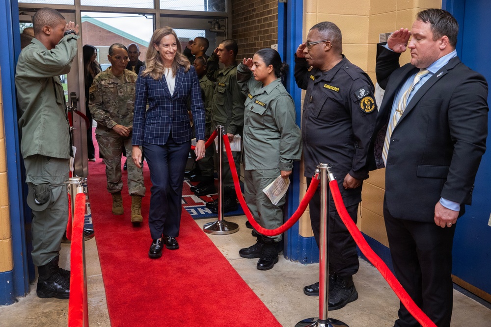 A formally dressed woman in a navy blue plaid blazer walks along a red carpet through what appears to be a military or law enforcement facility. She is being received with salutes from uniformed personnel on both sides, suggesting she is a dignitary or official visitor. The setting includes red rope barriers, brick walls, and a mix of military and civilian personnel.