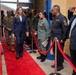 A formally dressed woman in a navy blue plaid blazer walks along a red carpet through what appears to be a military or law enforcement facility. She is being received with salutes from uniformed personnel on both sides, suggesting she is a dignitary or official visitor. The setting includes red rope barriers, brick walls, and a mix of military and civilian personnel.