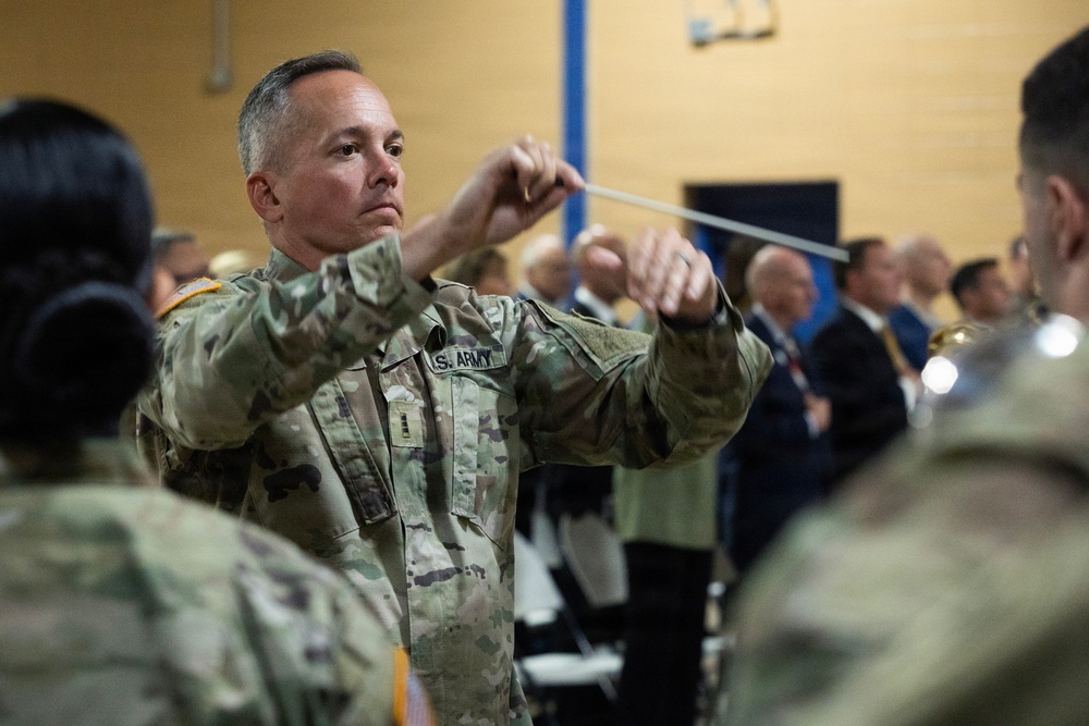 A U.S. Army soldier in OCP (Operational Camouflage Pattern) uniform is conducting a military band during what appears to be a formal ceremony or event. The conductor is holding a baton with both hands raised, directing musicians in uniform. An audience of civilians and military personnel is visible in the background.