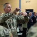 A U.S. Army soldier in OCP (Operational Camouflage Pattern) uniform is conducting a military band during what appears to be a formal ceremony or event. The conductor is holding a baton with both hands raised, directing musicians in uniform. An audience of civilians and military personnel is visible in the background.