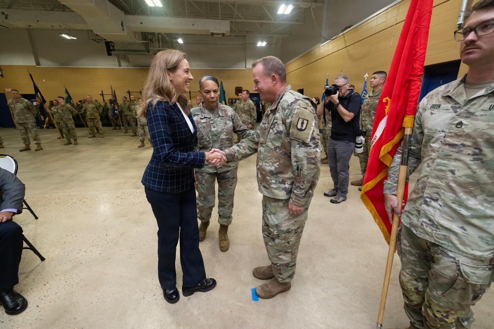 A civilian woman in a navy blue blazer is shaking hands with a senior military officer in Army combat uniform during what appears to be a formal military ceremony. The event is taking place in a large indoor gymnasium or drill hall with numerous uniformed soldiers standing in formation in the background. A soldier holding a red unit guidon flag stands to the right of the frame.