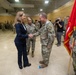 A civilian woman in a navy blue blazer is shaking hands with a senior military officer in Army combat uniform during what appears to be a formal military ceremony. The event is taking place in a large indoor gymnasium or drill hall with numerous uniformed soldiers standing in formation in the background. A soldier holding a red unit guidon flag stands to the right of the frame.
