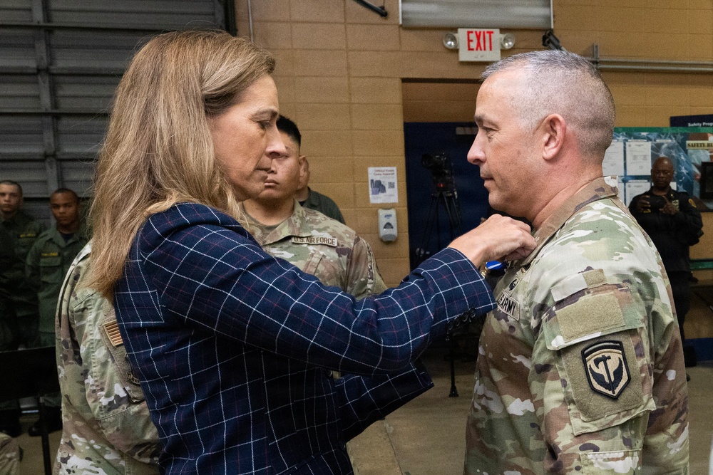 A woman in civilian attire (blue plaid blazer) is pinning a medal or insignia onto a U.S. Army soldier in OCP (Operational Camouflage Pattern) uniform during what appears to be a military awards or promotion ceremony. The event is taking place indoors, likely in a military facility or gymnasium, with several other military personnel observing in the background. The soldier receiving the pin wears a unit patch on his left shoulder consistent with U.S. Army affiliation.