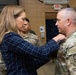A woman in civilian attire (blue plaid blazer) is pinning a medal or insignia onto a U.S. Army soldier in OCP (Operational Camouflage Pattern) uniform during what appears to be a military awards or promotion ceremony. The event is taking place indoors, likely in a military facility or gymnasium, with several other military personnel observing in the background. The soldier receiving the pin wears a unit patch on his left shoulder consistent with U.S. Army affiliation.