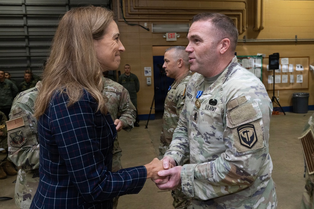 A civilian woman in a navy plaid blazer is shaking hands with a U.S. Army soldier in OCP (Operational Camouflage Pattern) uniform during what appears to be a military ceremony or official visit. The soldier is wearing a medal on his chest, suggesting a decoration or award ceremony has taken place. The setting is an indoor military facility, likely a hangar or large bay, with additional military personnel visible in the background.
