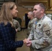 A civilian woman in a navy plaid blazer is shaking hands with a U.S. Army soldier in OCP (Operational Camouflage Pattern) uniform during what appears to be a military ceremony or official visit. The soldier is wearing a medal on his chest, suggesting a decoration or award ceremony has taken place. The setting is an indoor military facility, likely a hangar or large bay, with additional military personnel visible in the background.