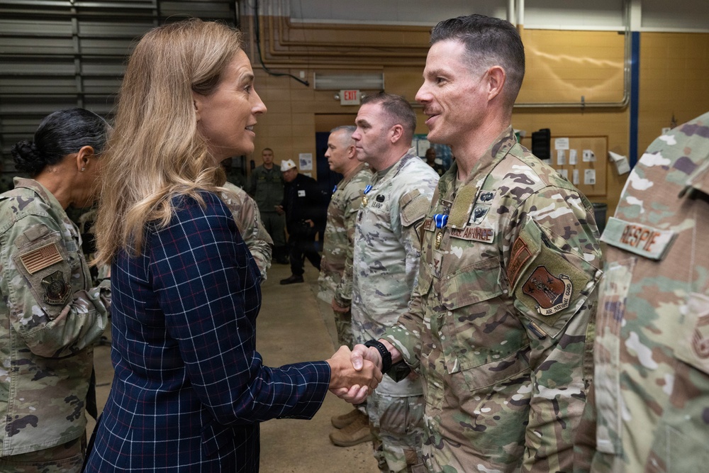 A civilian woman in a navy blue plaid blazer is shaking hands with a U.S. Air Force military officer in OCP (Operational Camouflage Pattern) uniform. The setting appears to be inside a military hangar or facility, with several other uniformed Air National Guard personnel standing in the background. The interaction appears to be a formal greeting or recognition ceremony.
