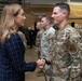 A civilian woman in a navy blue plaid blazer is shaking hands with a U.S. Air Force military officer in OCP (Operational Camouflage Pattern) uniform. The setting appears to be inside a military hangar or facility, with several other uniformed Air National Guard personnel standing in the background. The interaction appears to be a formal greeting or recognition ceremony.