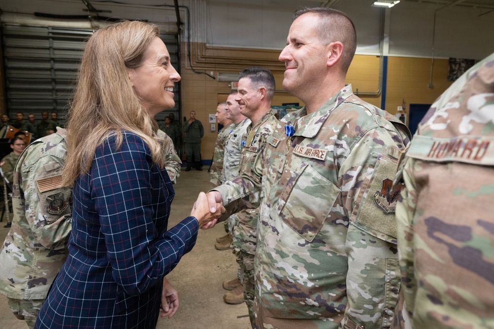A civilian woman in a navy blue plaid dress is shaking hands with a U.S. Air Force service member wearing OCP (Operational Camouflage Pattern) uniform and a medal. The setting appears to be an indoor military facility or hangar, with additional uniformed personnel lined up in the background. The interaction suggests a formal recognition or greeting ceremony.