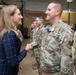 A civilian woman in a navy blue plaid dress is shaking hands with a U.S. Air Force service member wearing OCP (Operational Camouflage Pattern) uniform and a medal. The setting appears to be an indoor military facility or hangar, with additional uniformed personnel lined up in the background. The interaction suggests a formal recognition or greeting ceremony.