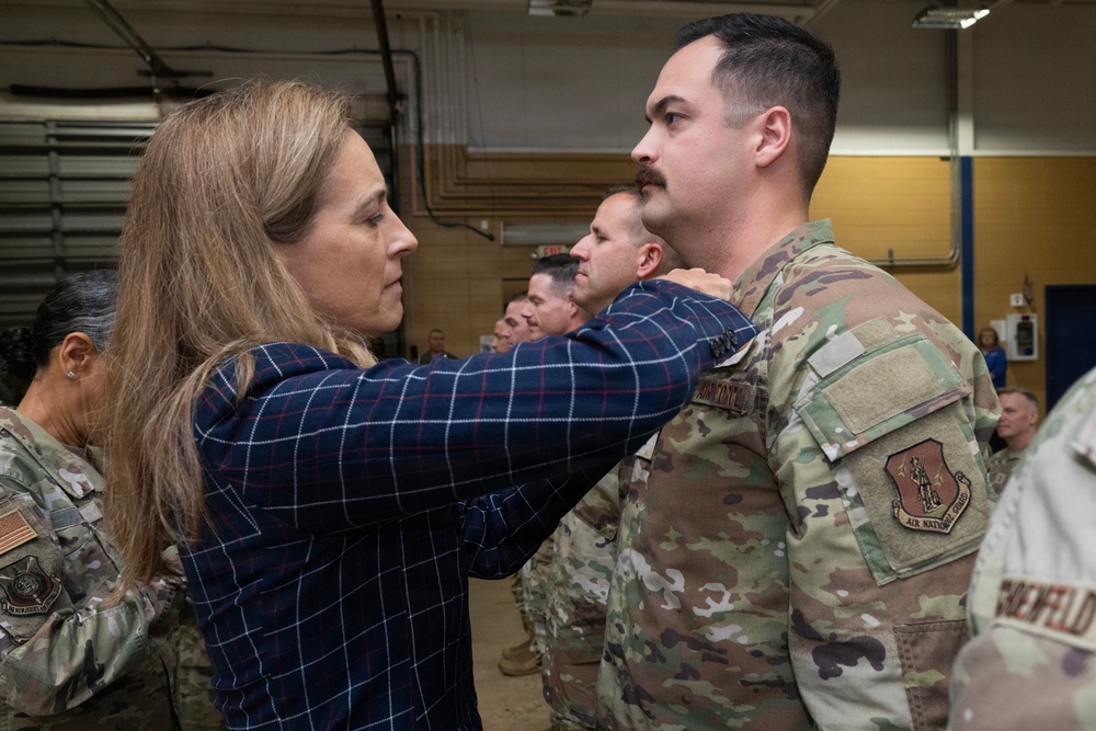 A civilian woman in a plaid shirt is pinning something onto the collar or shoulder area of a male service member in OCP (Operational Camouflage Pattern) uniform. This appears to be a military promotion or award ceremony taking place inside a hangar or maintenance bay. Several other uniformed personnel are visible in the background observing the ceremony.