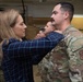 A civilian woman in a plaid shirt is pinning something onto the collar or shoulder area of a male service member in OCP (Operational Camouflage Pattern) uniform. This appears to be a military promotion or award ceremony taking place inside a hangar or maintenance bay. Several other uniformed personnel are visible in the background observing the ceremony.
