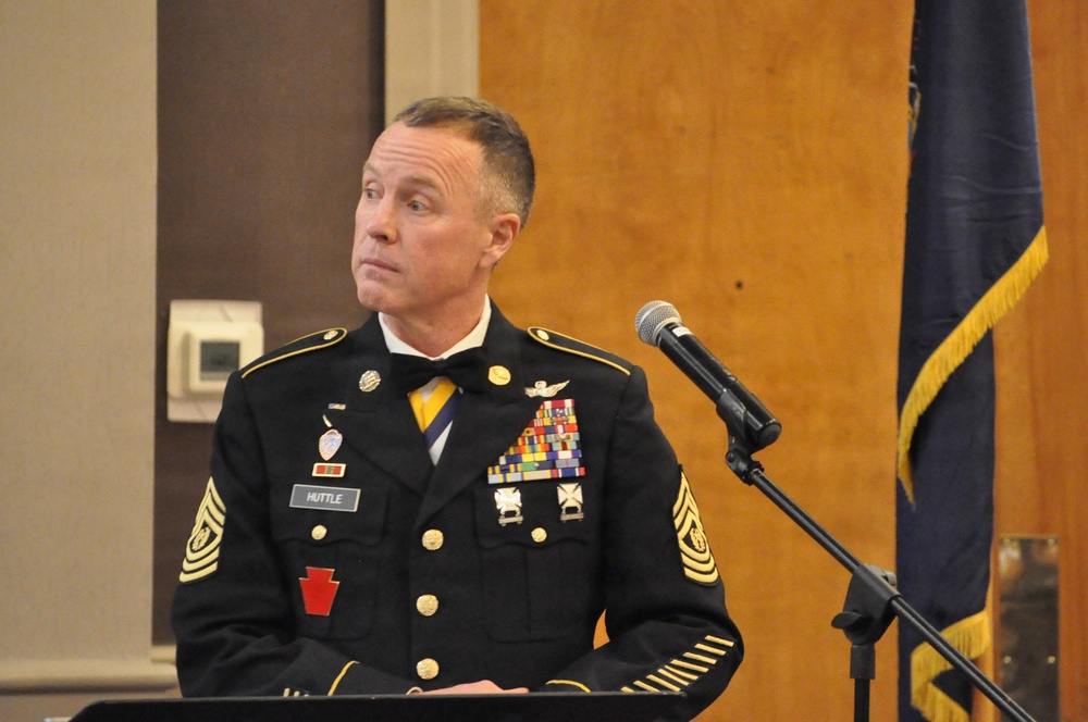 A U.S. Army senior non-commissioned officer identified by nameplate as 'HUTTLE' stands at a podium with a microphone, wearing the Army Service Uniform (ASU) in dress black. He displays the rank insignia of a Sergeant Major or Command Sergeant Major on his sleeves, along with an extensive array of service ribbons, badges, and unit patches. A dark blue flag with gold fringe is visible in the background, consistent with a formal military ceremony or official speaking engagement.