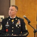 A U.S. Army senior non-commissioned officer identified by nameplate as 'HUTTLE' stands at a podium with a microphone, wearing the Army Service Uniform (ASU) in dress black. He displays the rank insignia of a Sergeant Major or Command Sergeant Major on his sleeves, along with an extensive array of service ribbons, badges, and unit patches. A dark blue flag with gold fringe is visible in the background, consistent with a formal military ceremony or official speaking engagement.