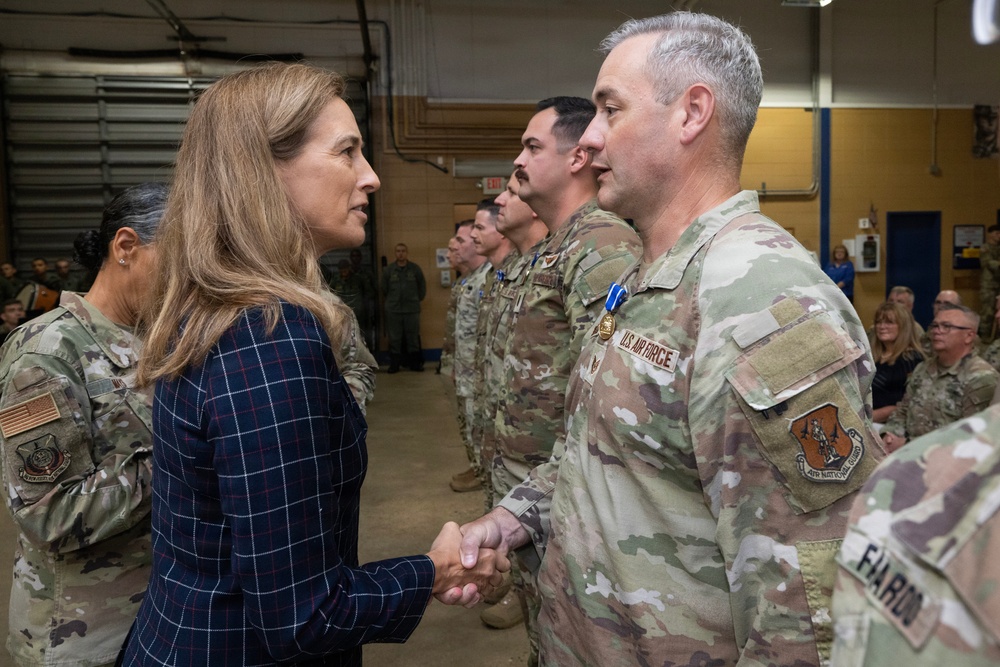 A civilian woman in a navy plaid blazer is shaking hands with a U.S. Air Force service member in OCP (Operational Camouflage Pattern) uniform during what appears to be a formal military ceremony. The service member is wearing a medal, suggesting this is an awards or recognition event. Multiple uniformed military personnel are standing in formation in the background inside what appears to be a hangar or large military facility.
