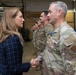 A civilian woman in a navy plaid blazer is shaking hands with a U.S. Air Force service member in OCP (Operational Camouflage Pattern) uniform during what appears to be a formal military ceremony. The service member is wearing a medal, suggesting this is an awards or recognition event. Multiple uniformed military personnel are standing in formation in the background inside what appears to be a hangar or large military facility.