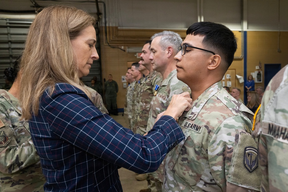 A civilian woman in a blue plaid blazer is pinning a medal or award onto a U.S. Army soldier in OCP (Operational Camouflage Pattern) uniform during a formal military awards ceremony. Several other military personnel in various uniforms are lined up in the background, some already bearing medals on their chests. The event appears to be taking place inside a military facility or hangar.