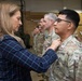 A civilian woman in a blue plaid blazer is pinning a medal or award onto a U.S. Army soldier in OCP (Operational Camouflage Pattern) uniform during a formal military awards ceremony. Several other military personnel in various uniforms are lined up in the background, some already bearing medals on their chests. The event appears to be taking place inside a military facility or hangar.