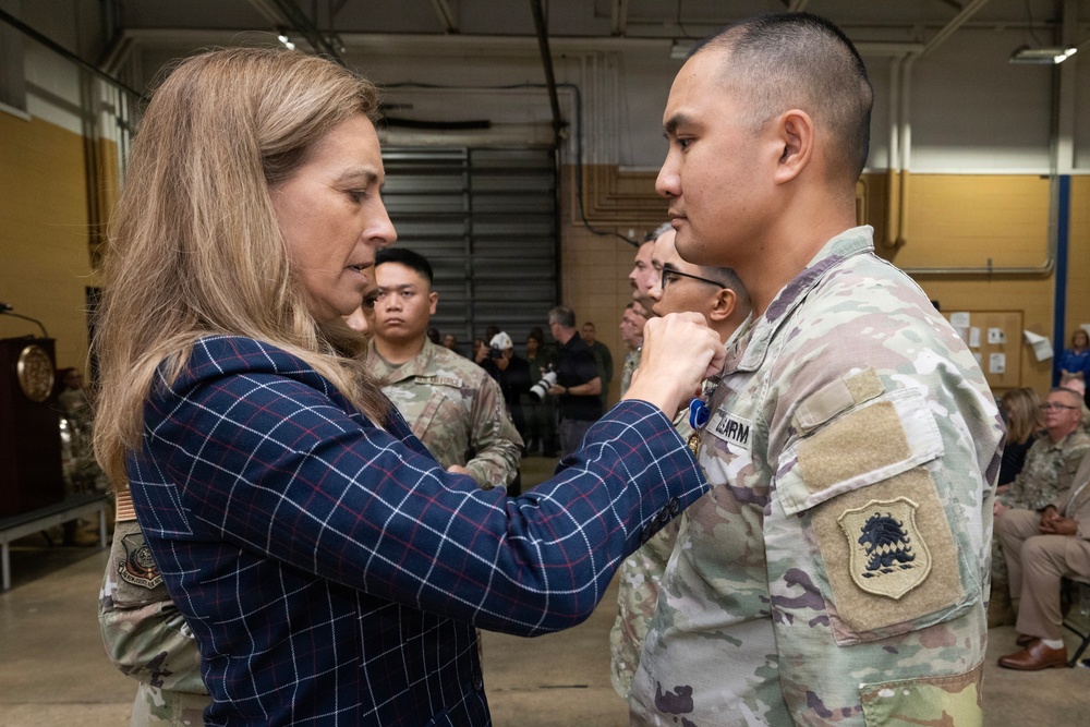 A woman in civilian attire is pinning a medal onto a soldier in U.S. Army OCP (Operational Camouflage Pattern) uniform during what appears to be a formal military awards ceremony. The event is taking place inside a military facility or hangar, with other uniformed personnel and civilians visible in the background. The ceremony appears to be an official recognition or decoration event.